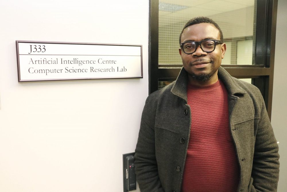 Assistant Professor of Computer Science Blessing Ogbuokiri stands next to a sign saying “J333. Artificial Intelligence Centre Computer Science Research Lab” with a window framing him in the background.