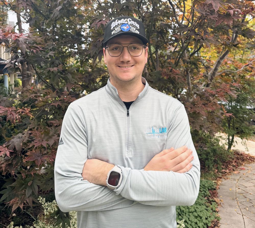 A man in a baseball cap poses outside in front of a garden.