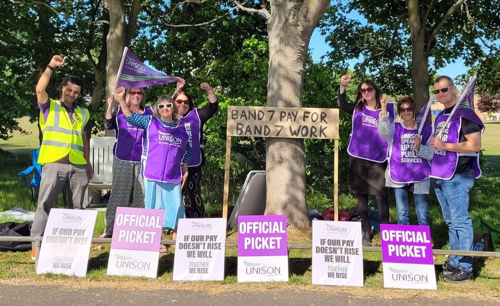 Pickets stand holding flags by a sign demanding band 7 pay for band 7 work