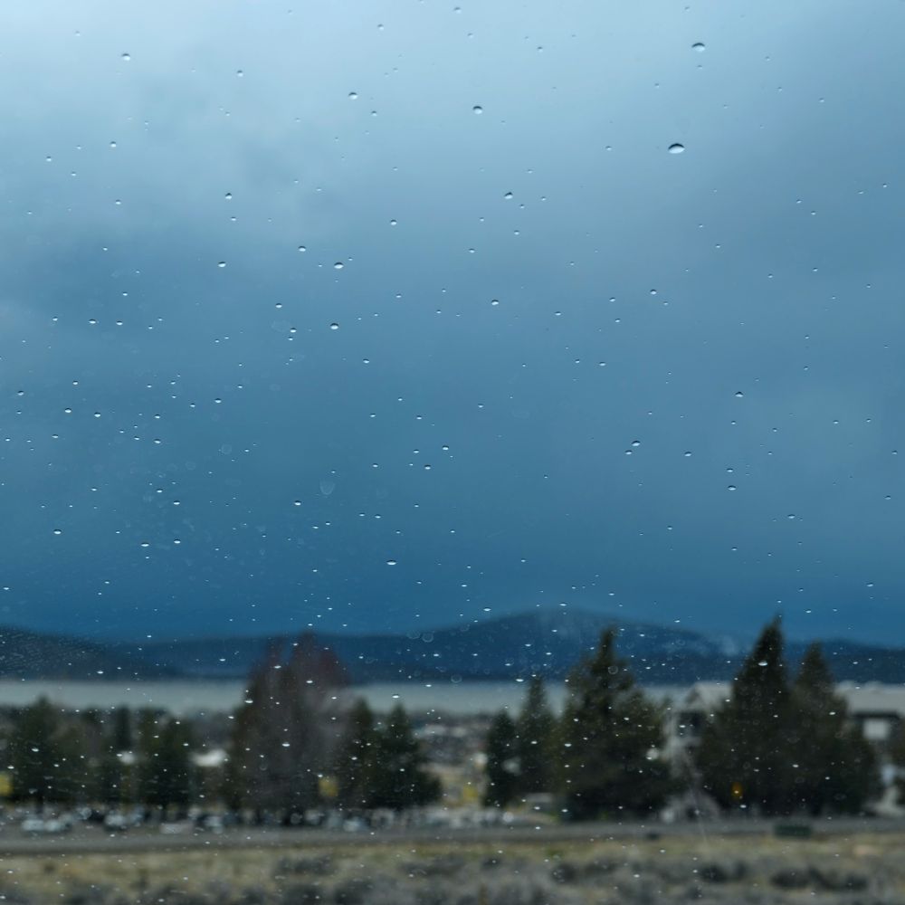 looking out of the car window toward the Oregon Technical Institute
from a favorite hiking hill. There are a line of trees with a indistinct lake and mountains in the background. There are faint raindrops on the windshield.