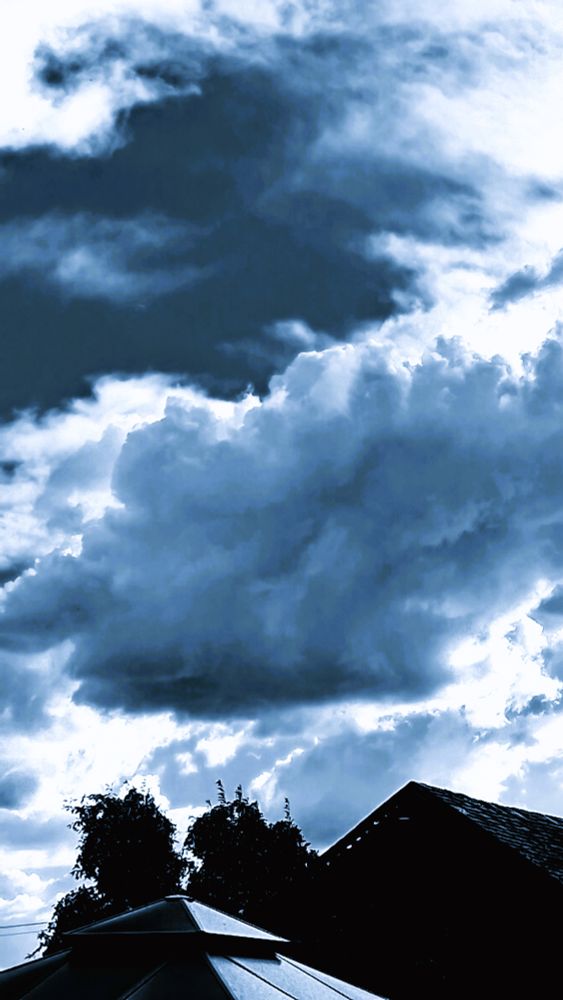puffy gray blue clouds above two rooftops and two trees.
