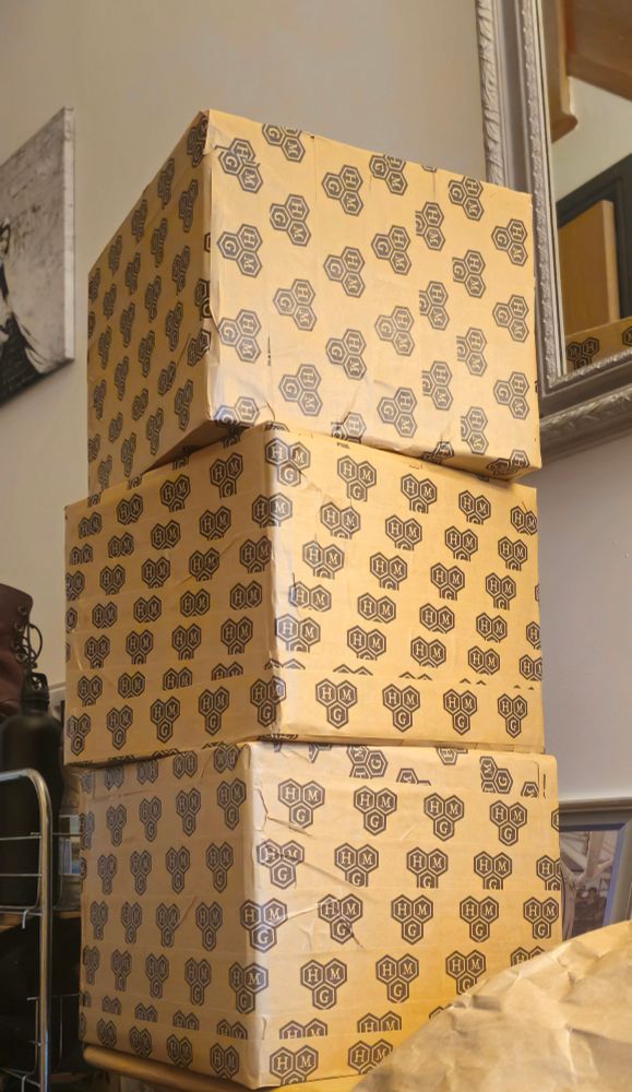 Three boxes stacked on each other by the front door of a home. They're around 32inches square, and wrapped entirely in Hive Mind Games branded tape.