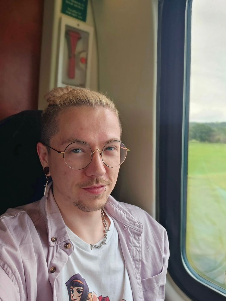 Photo of me on a train. I'm wearing a white Cowboy Bebop tee with a ping over shirt. I've got round glasses, and bleached hair with the roots showing tied back on a bun. 