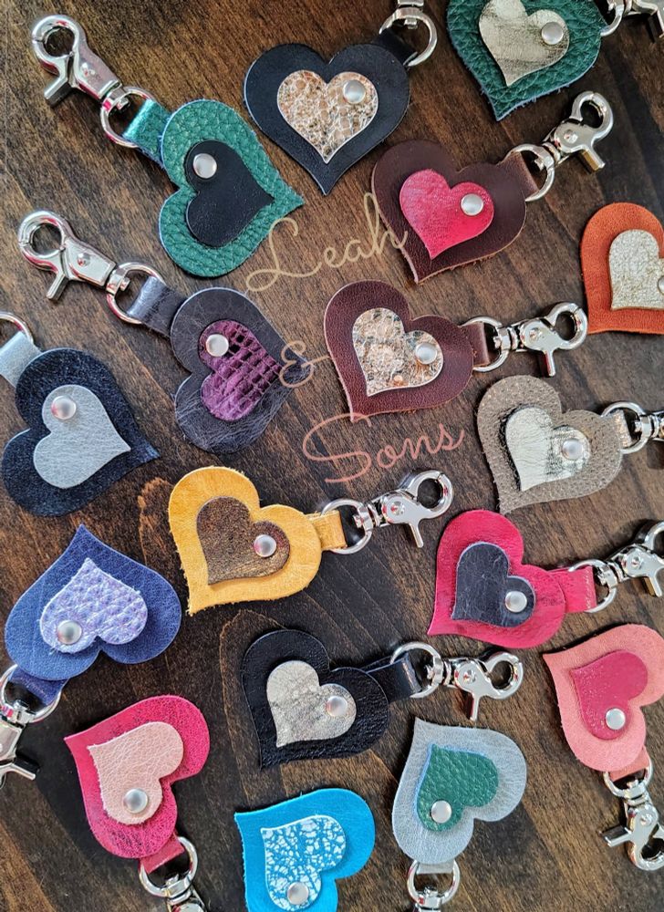 Colorful leather heart charms with various metallic leathers, all with silver trigger clasps, and "Leah & Sons" lightly written across the picture.