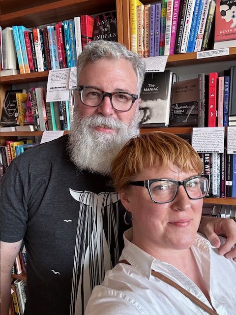 A handsome bearded, bespectacled man smiles as he places his arm on the shoulder of a beautiful, short-haired redheaded woman also wearing glasses. He wears a dark blue T-shirt with a white image of raised whale flukes while she sports a crisp, collared white shirt open at the neck. Both stand in front of a set of shelves containing countless books and several shelf talkers recommending same.