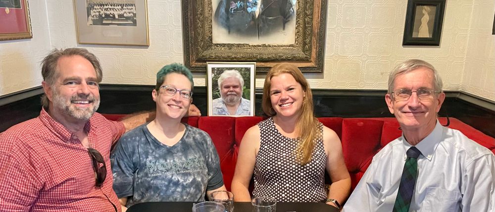 Four people (L to R: Mark, me, Stephanie, and Rob) in a pub booth with a picture of a different man (Simon, RIP) centered behind them. 