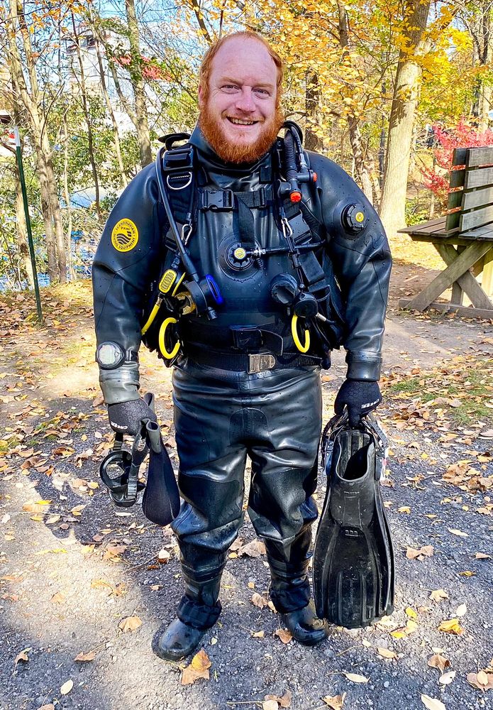 A photo of human Verne standing in his dive gear with a lake in the background behind several trees. He's wearing a black rubber drysuit, BCD and gloves, and is holding a pair of black fins, a black mask and a hood in his hands.