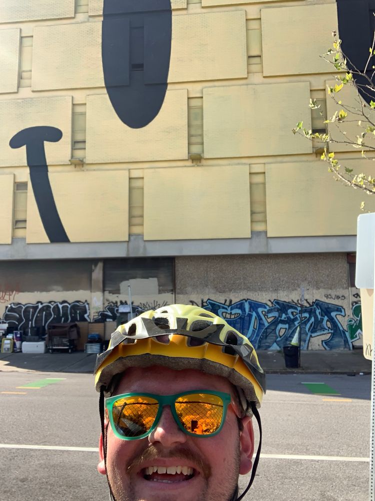 Grinning selfie of a person wearing a yellow bike helmet and sunglasses, standing in front of a building with a giant yellow smiley face.