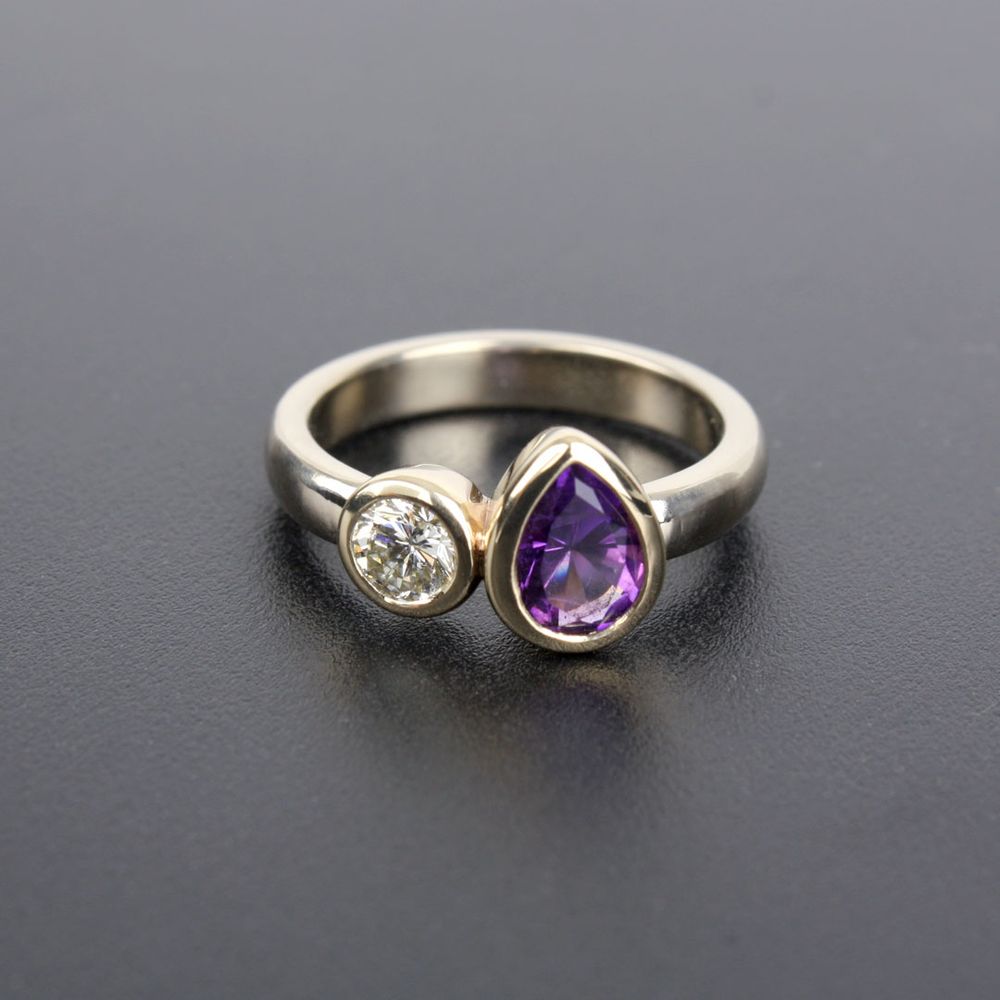 An engagement ring with a sparkling round diamond and a pear-shaped deep purple amethyst. The setting is yellow gold, with a palladium white gold band. The ring sits on a dark background.