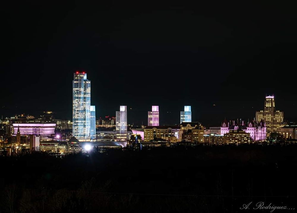 a photograph by A. Rodriguez of the Albany skyline. the state offices are illuminated in blue, pink, and white lights. 