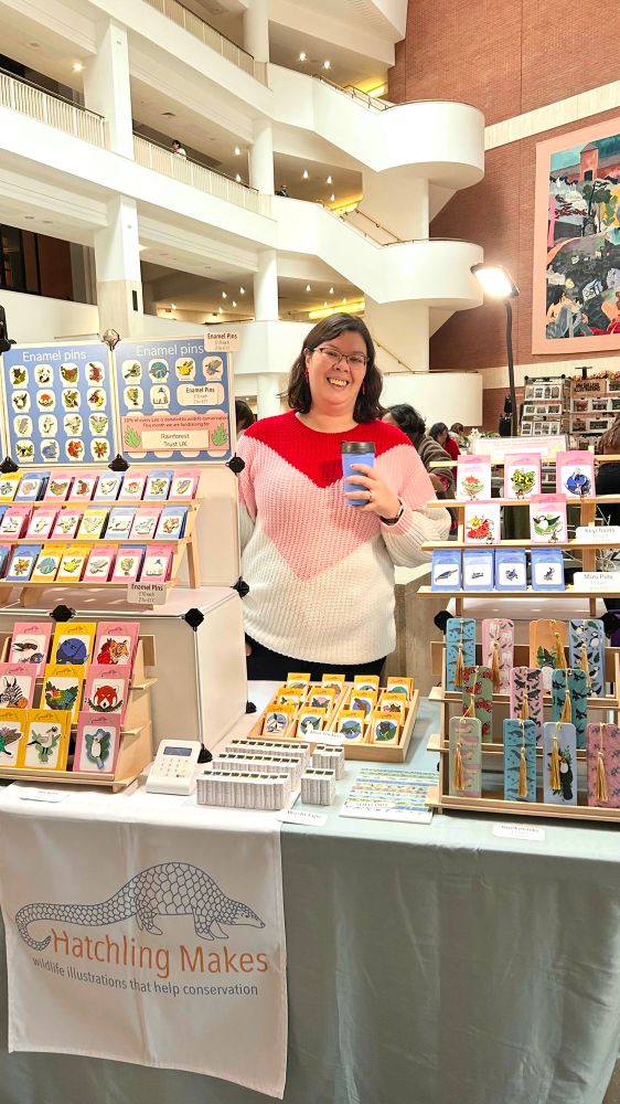 A photo of Tash, a white passing woman with dark hair and glasses, standing behind a market table set up with products from her small business