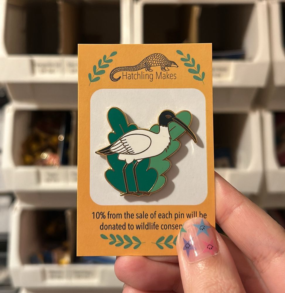 A hand is holding an enamel pin of a pin chicken on a yellow and white backing card