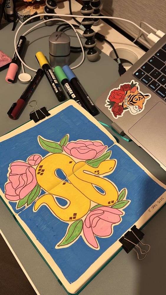 An open sketchbook with a yellow snake and 3 pink flowers illustrated across two pages on a blue background 