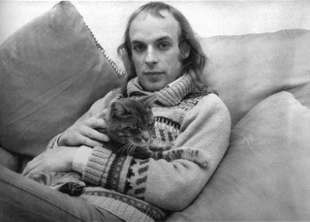 Brian Eno, mid-1970s, sitting in a pile of pillows wearing a Nordic-style sweater and holding a tabby cat.