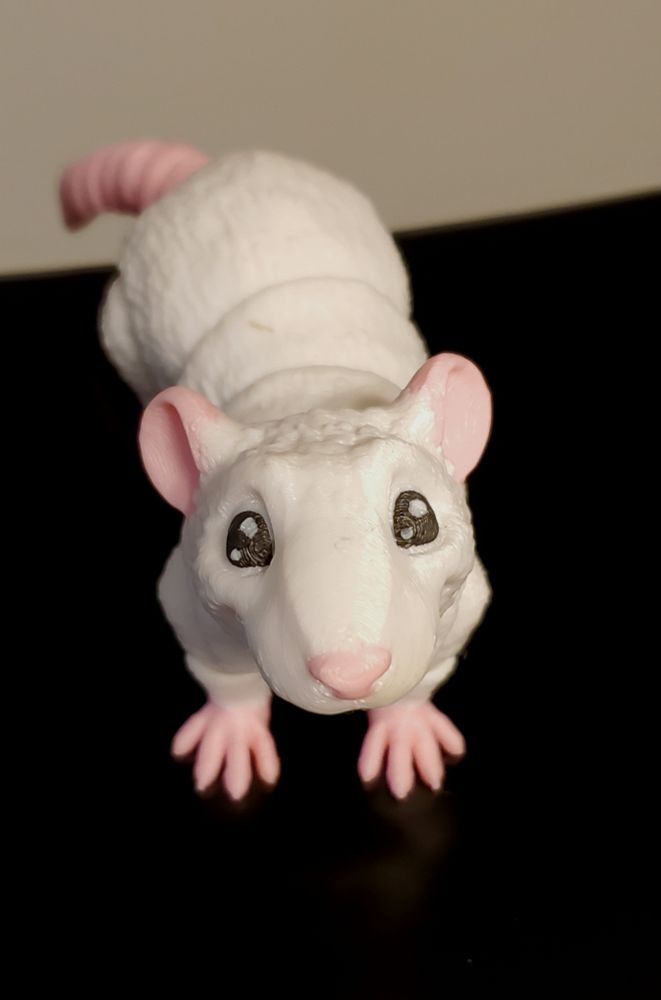A white articulated 3D printed rat posed to look up at the camera. It's feet, nose, ears, and tail are pink. It's standing on a black base against a grey background. 