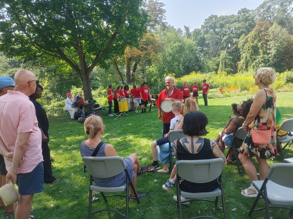 Folks standing and sitting in folding chairs in the park listening to Samba band leader