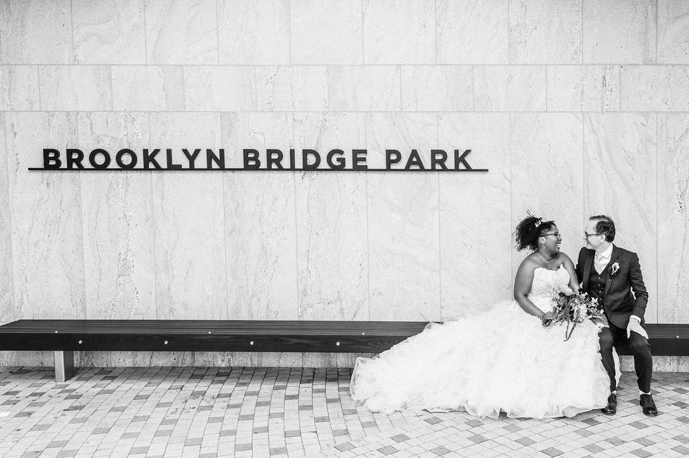black and white photo of me and The Husband on our wedding day, sitting a long bench, towards the right end. behind us to the left is a sign reading BROOKLYN BRIDGE PARK. we are looking at each other and smiling; he's lookin all sharp in his suit, and I am looking all princessy wearing a strapless wedding gown with an enormous train fully laid out and a tiara, and holding a bouquet