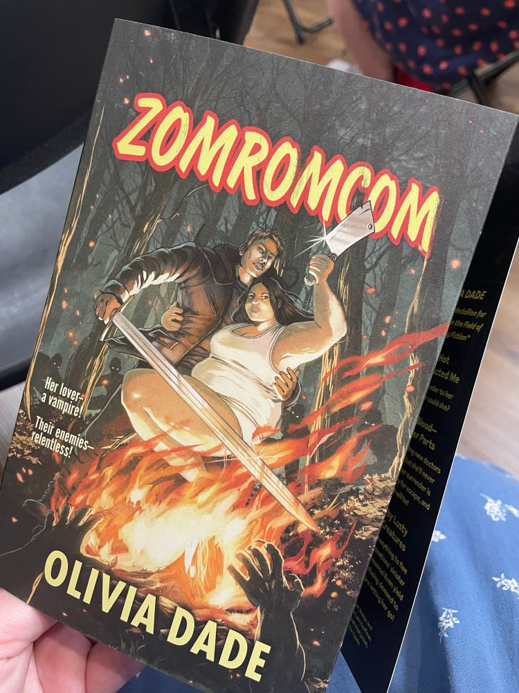 Photo of a promotional dusk jacket for Olivia Dade’s book Zomromcom. The cover illustration features a man and a woman, both armed with sharp implements, facing some shadowy creatures across a bonfire; the art absolutely rules. 