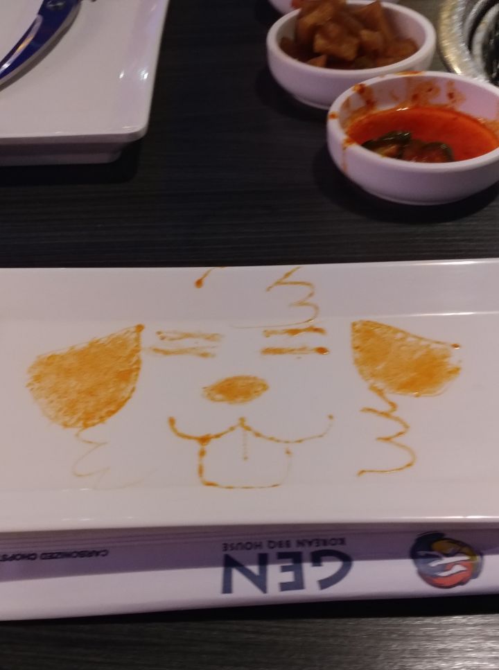 A dog drawn using condiments on a plate on the edge of a KBBQ table. Banchon is present in the photo and a chopsticker wrapper that says "Gen".