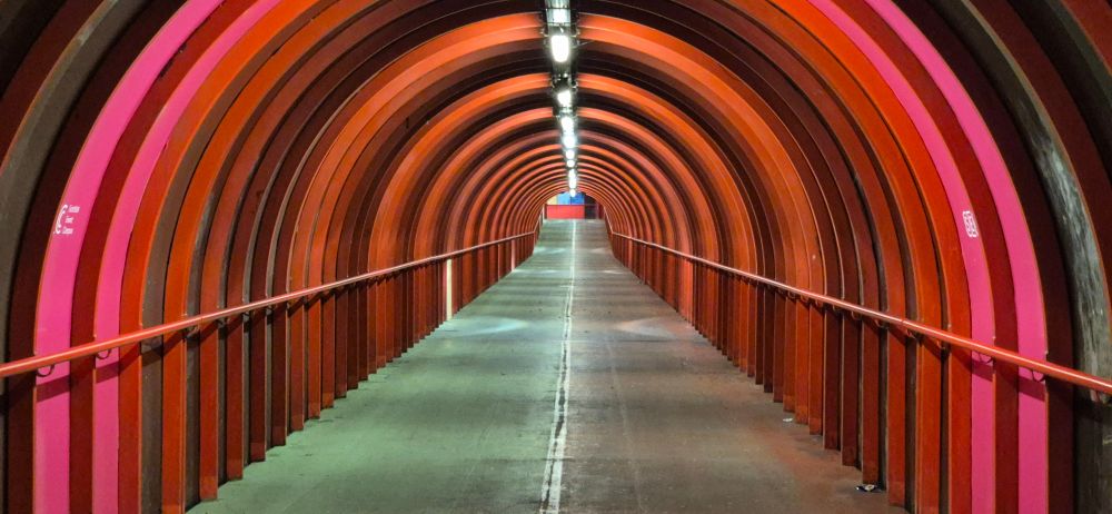 An empty footbridge tunnel disappears into the distance, with overhead lights creating dark and light stripes on the red metalwork.