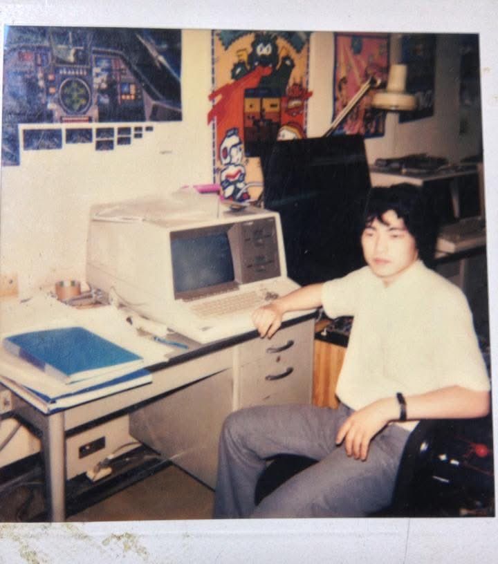 A man with a white shirt and grey pants in front of a white computer. On the wall: several game posters.