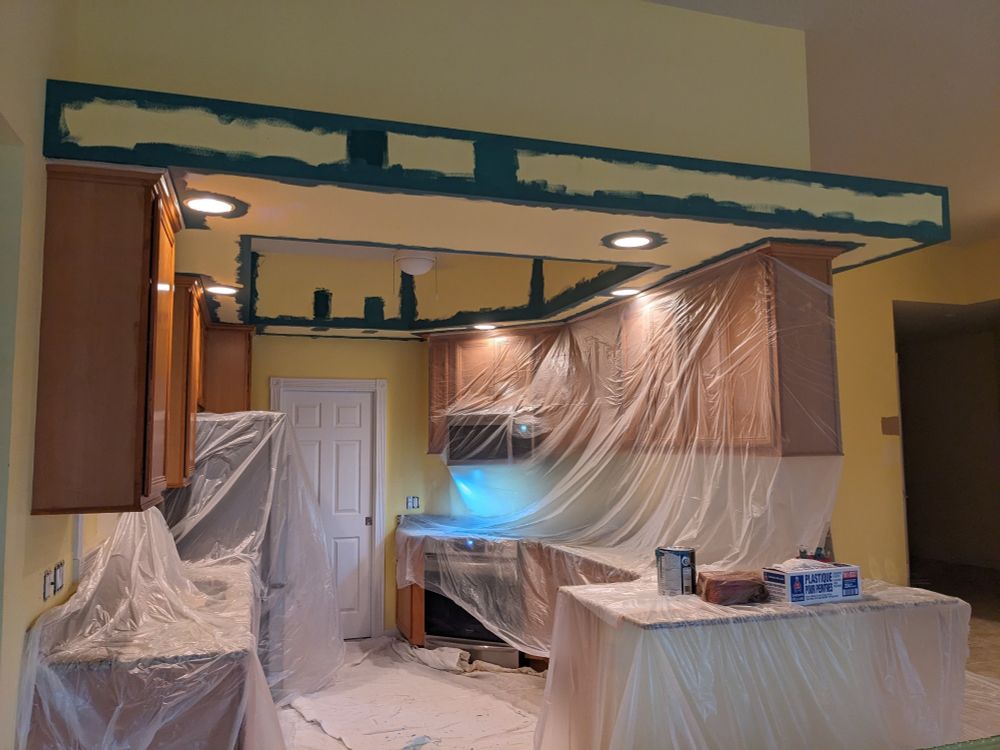 A head-on photo of a yellow kitchen, taken from an open-concept entryway. There are plastic tarps covering most of the hanging cabinets, countertop, and floors. The lights are on overhead as there is no natural light source. The lowest portion of the ceiling and upper walls have been edged with teal paint.