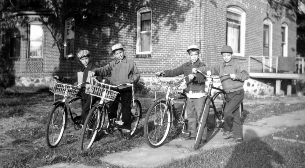 Four boys between the ages of 10 and 7 astride their balloon tire bicycles. A brick house is in the background.