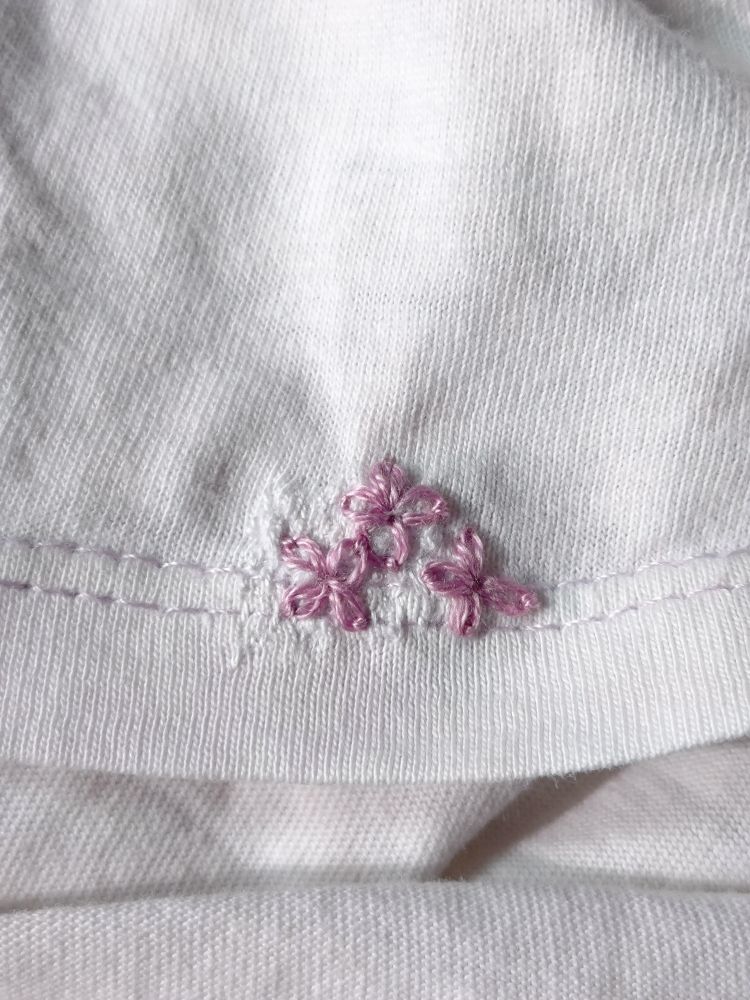 The hem of a white t-shirt, darned with white thread, and embroidered with three lilac coloured lazy daisy flowers.