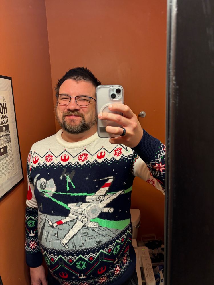 A man stands in an orange bathroom taking a mirror selfie in a Star Wars Death Star trench fight scene “holiday” sweater.
