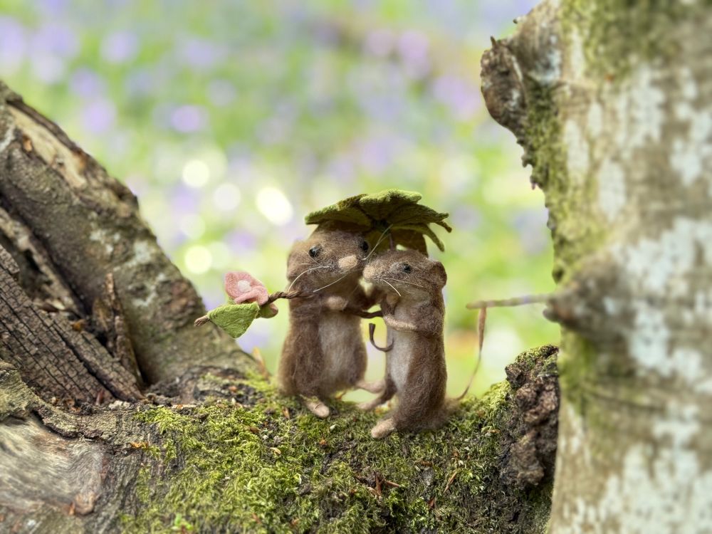 Two felted mice sheltering under a felt stitched leaf umbrella in a bluebell wood 