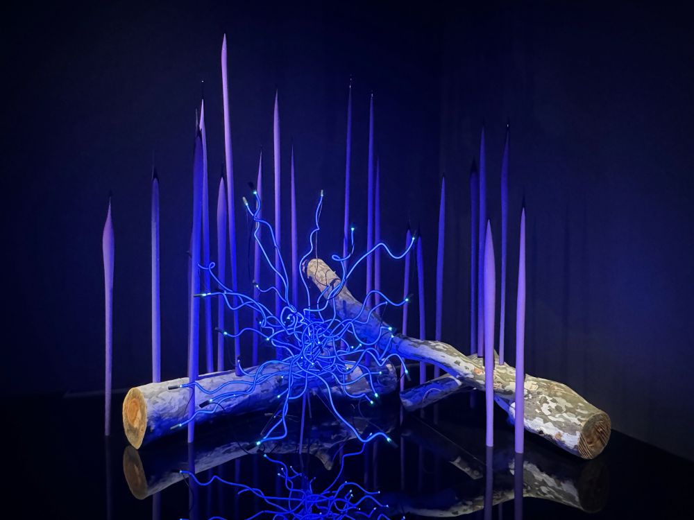 Photo of a violet neon “tumbleweed” with violet glass reeds and tree trunks surrounding it