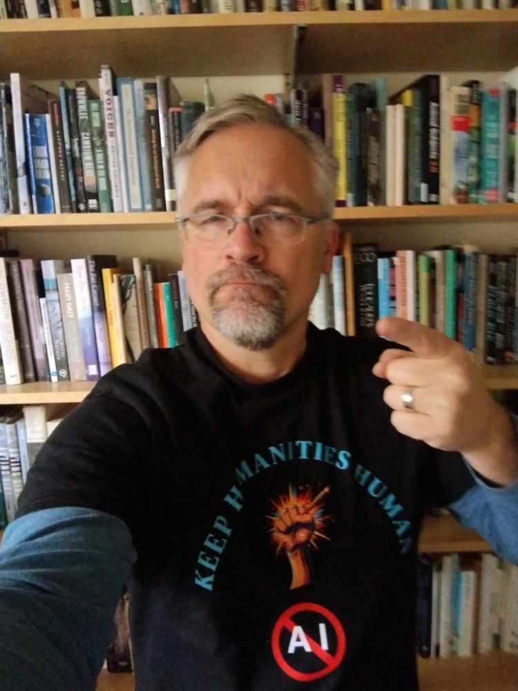 Your humble correspondent, looking grumpy and wearing a t-shirt that reads "Keep Humanities Human." It also has a raised fist holding a pencil, and the letters A and I inside a circle with a line through it.