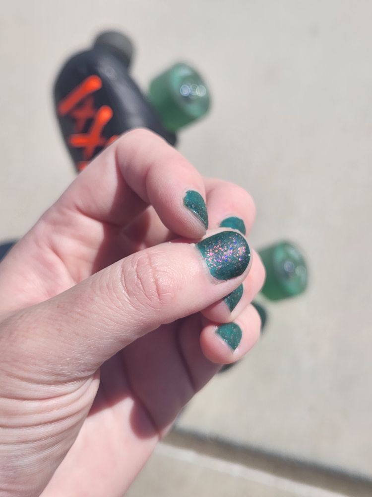 A person's hand with dark teal sparkly nail polish and a skate with teal wheels in the background 