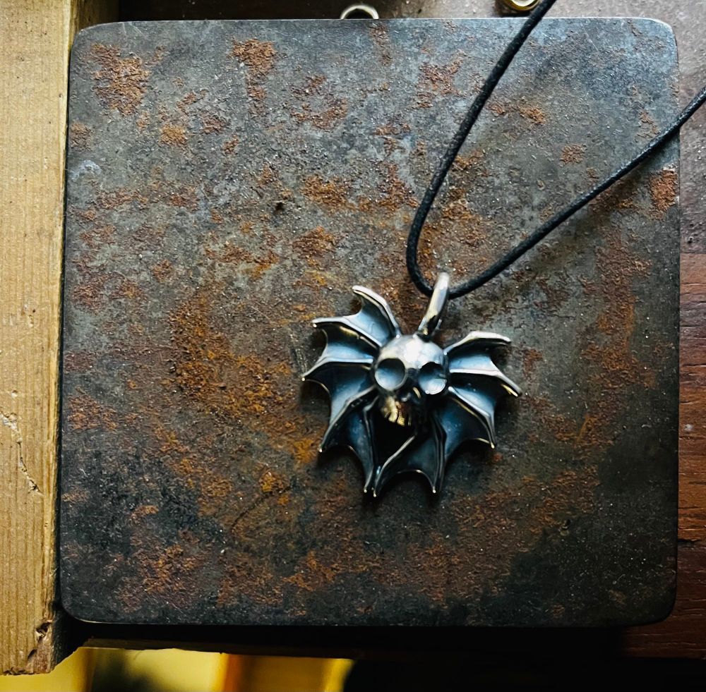 A silver pendant with a crude  skull in the centre and bat wings is photographed on a rusty block of metal. The red rust contrasts with the oxidised surface of the silver