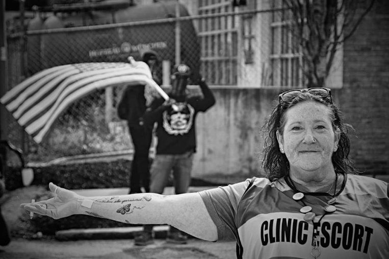 A woman wearing a clinic escort vest is giving you a Vanna White impression to show you the Proud Boys in the background. Her tattoo says “Nevertheless, she persisted” in her mother’s handwriting. She is very proud of herself and it shows.