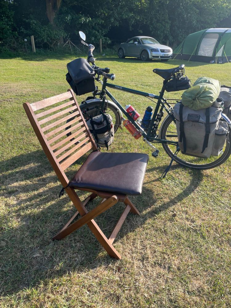 A parked, fully loaded touring bike with a folding wooden chair next to it.
