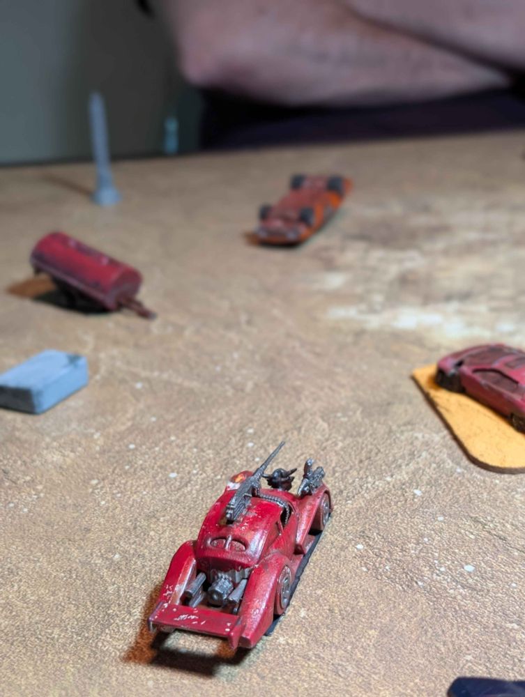 In focus in the foreground a red performance car with a .50 calibre machine gun mounted on the roof sits triumphant on the sandy arena floor. Out of focus in the background sits the flipped corpse of an orange muscle car. Assorted pieces of wreckage are strewn around the battleground.