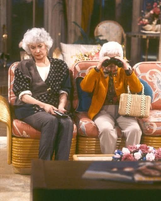 Dorothy and Sophia sitting on the living room couch. Dorothy looks annoyed, and Sophia is ĺoking through a pair of binoculars. 