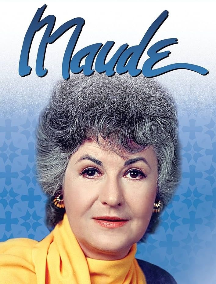 A photo of Bea Arthur in gold earrings and a yellow scarf, with the word "Maude" written above her 