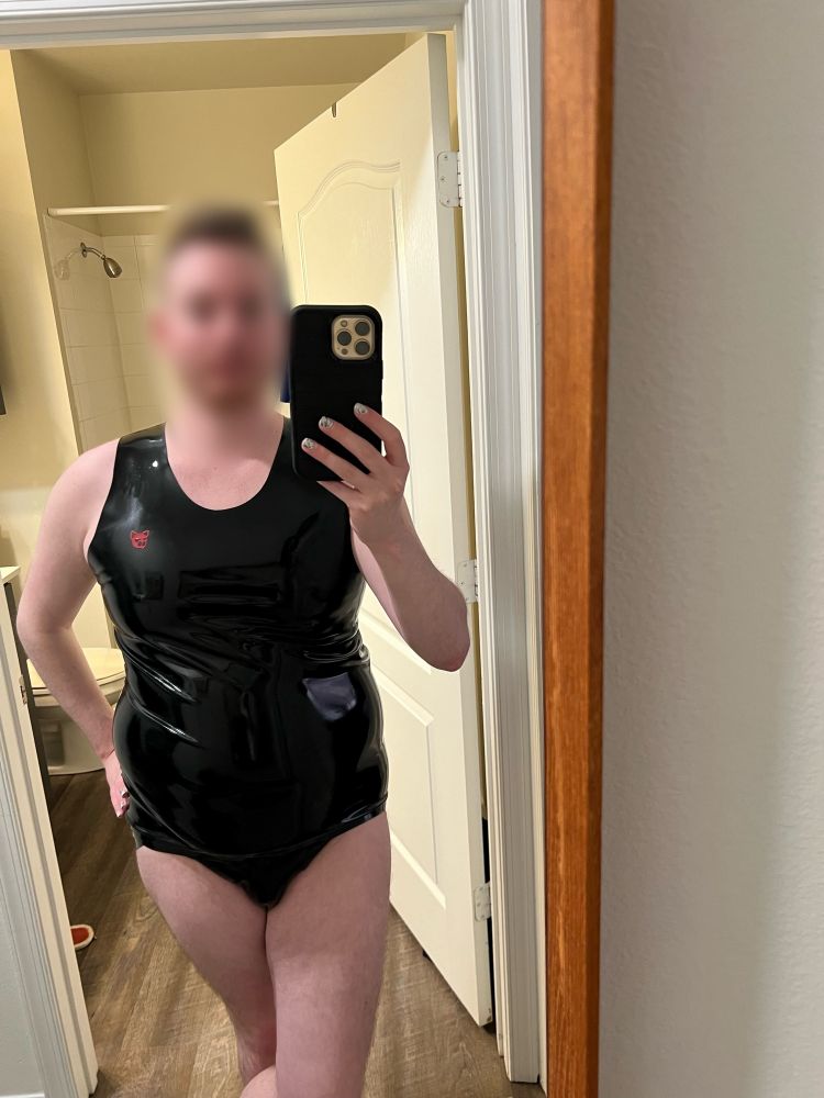 Mirror selfie of a white human with a blurred face, wearing black rubber tank and black rubber jock strap. They are holding a phone in a black case and is facing wooden framed mirror. Bathroom with tan walls and a white door is in the background.
