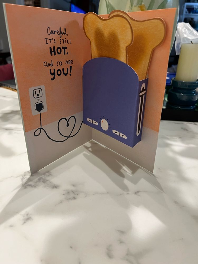 The inside of the card has an orange wall background with white wall socket on the left of the card. The bottom quarter of the card has a pale salmon background. Coming out of the wall socket is the plug meandering  and looped in the shape of heart at the bottom of the card. The plug leads to a pop out lavender toaster with two slices of golden toast popping out of the top of it. Each slice of toast has a slightly darkened heart, toasted into it. Written in black on the left side of a card in various fonts reads: “Careful it’s still HOT. And so are YOU!” The card is standing up right on a marble table. Above the card you can see part of a green floral vase. In the right of the card is a candle sitting in a turquoise, round candle holder.