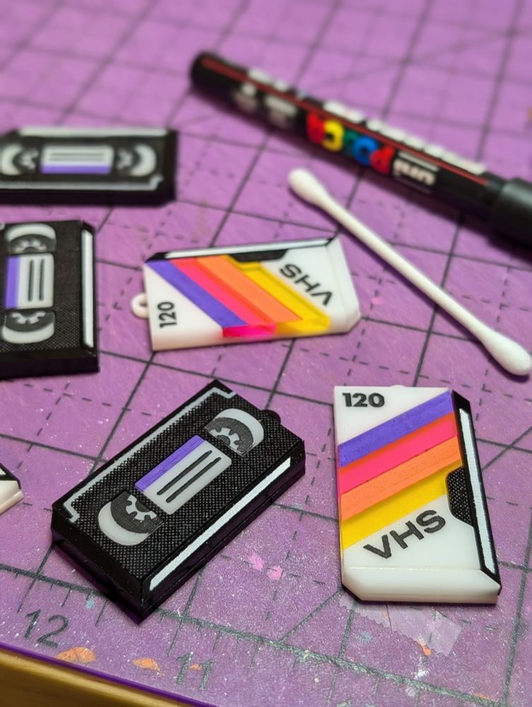 Photo of a workbench with handmade earring charms, a paint marker, and a cotton swab. Some of the charms look like a black VHS cassette with purple details, while the other charms look like the retro white and rainbow colored sleeve of an old blank VHS tape. 