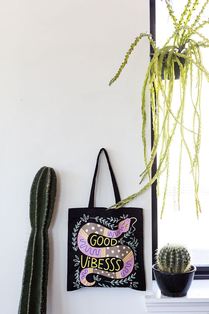 Lifestyle imagery of a black tote bag hanging on a wall surrounded by cacti. The tote bag features a snake illustration with the phrase "Good Vibesss". 