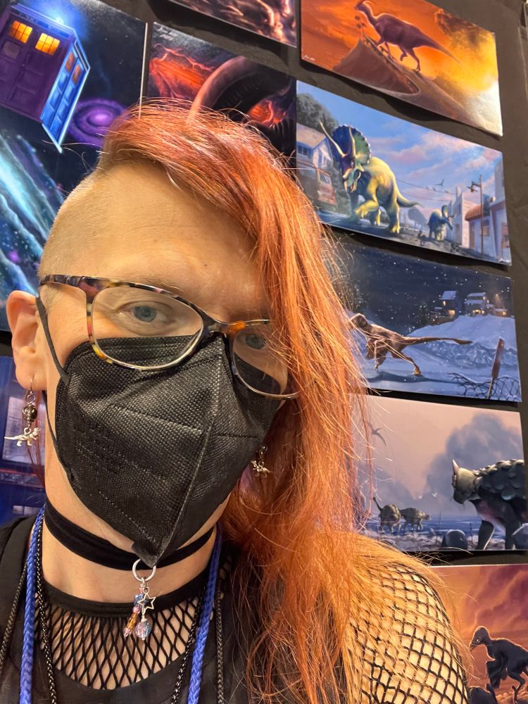 A picture of Ana standing in front of her dinosaur artwork. She is wearing a black face mask and dinosaur earrings.