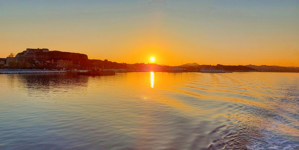 Sonnenuntergang über einer flache Küste mit Häusern und einer Festung links. Im Meer davor spiegelt sich die Sonne