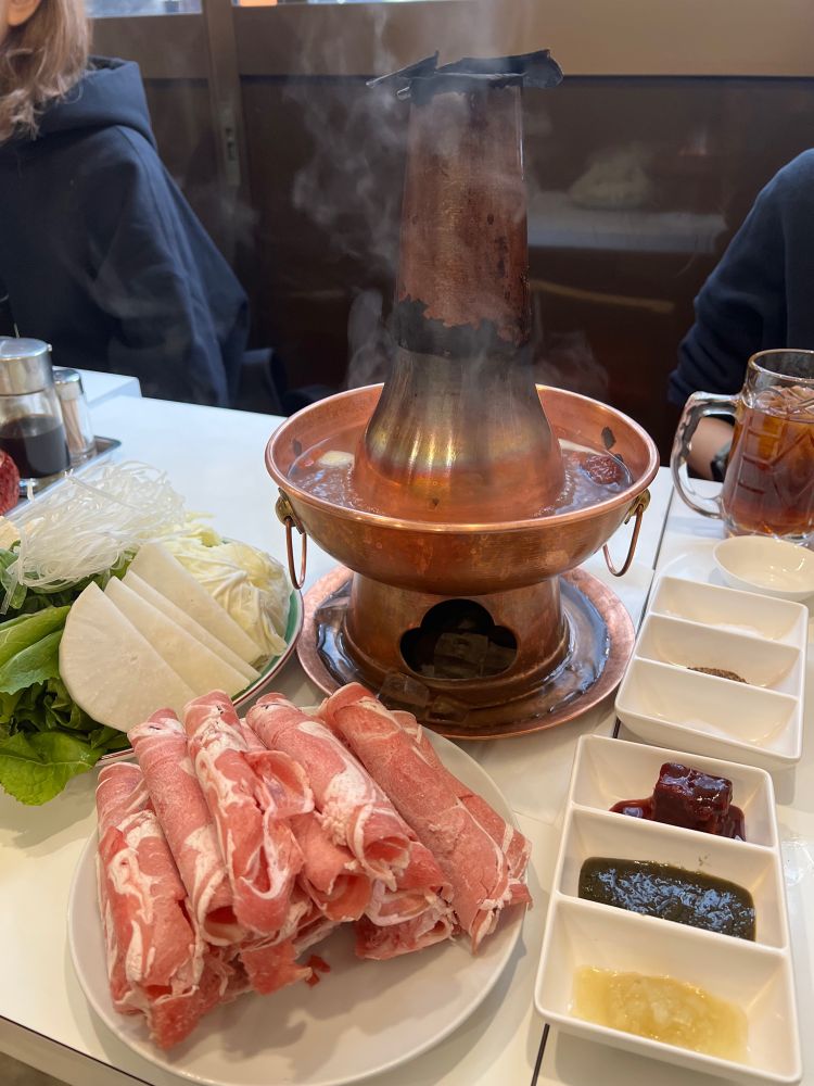 机の中央に置かれた火鍋子ホーコーツと呼ばれる煙突のある鍋から湯気が立っている。野菜と山盛りのラム肉の横に、飲み物と6種類のタレが置いてある。