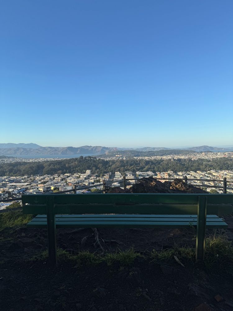 A photo from atop Grandview Park (aka Turtle Hill) in San Francisco showing a park bench and its view from atop the park of San Francisco, the Golden Gate Bridge, Marin county and beyond. There is not a single cloud in the sky. It's a beautiful day. 