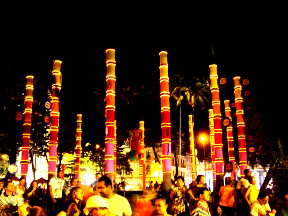 foto colorida noturna de colunas vermelhas e amarelas em círculo no meio de uma praça com muitas pessoas em volta