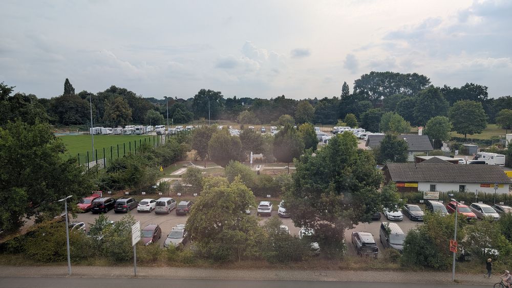 Im Vordergrund ein Parkplatz voller Autos. Links der Rand eines Fußballplatz. Hinter dem Parkplatz ein Minigolfplatz sowie ein Wohnmobilstellplatz. 