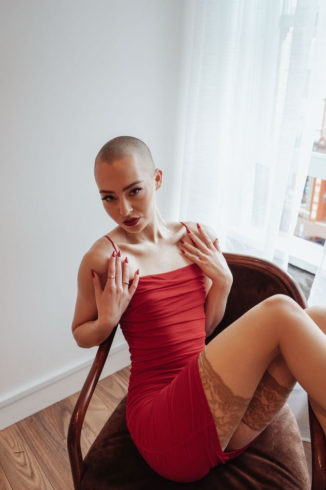 Darla sits sideways in a wooden chair with her legs draped over one of the armrests. She is wearing a red skintight dress and nude stockings. 